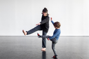 An adult and a child wearing athletic clothes kick out their feet in a dance studio