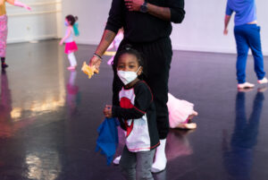 A young child wearing a spiderman shirt and face mask holds a colorful cloth, standing in front of an adult in a dance studio