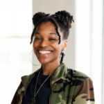 A Black woman wearing a camouflage jacket and her hair in two high buns smiles