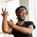 A Black man wearing athletic clothing holds an elegant dance pose with his arms, one crossing to the opposite shoulder while the other hand reaches with fingers splayed