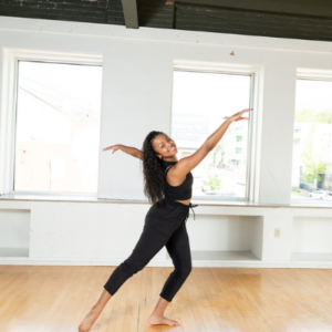 A Black woman in athletic clothes holds a ballet pose, with arms and leg outstretched, in a dance studio