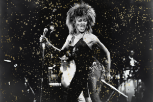 A black & white photograph of Tina Turner onstage, wearing a black dress. She is holding her microphone stand and singing passionately, looking off toward the side. Instruments are barely visible in the background, and there is a vintage-looking golden confetti-esque treatment over the image