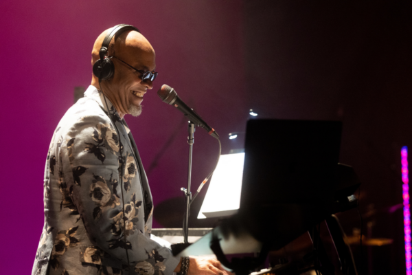 Dwayne Fulton is onstage playing the piano with a bright smile. He is a bald Black man, wearing sunglasses and headphones, dressed in a light blue suit jacket patterned with big white flowers. He is lit by an overhead stage light in front of a pink backdrop